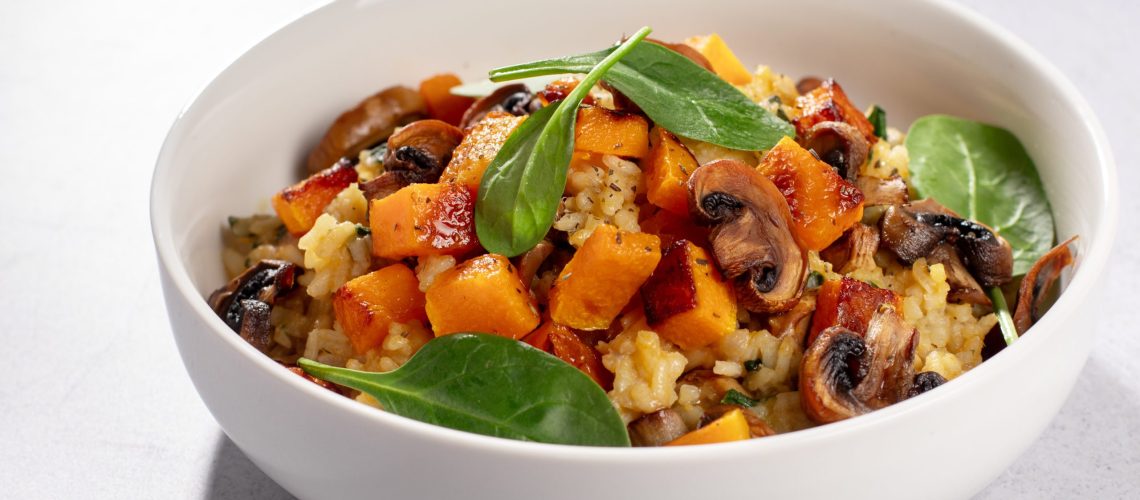 A vegan risotto with roasted pumpkin, mushrooms, and fresh spinach served in a white bowl on a light background, top view, closeup view